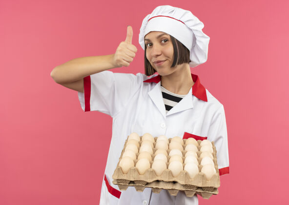 拿着身着厨师制服的年轻女厨师手捧一盒鸡蛋 在粉色背景上孤立地竖起大拇指制服厨师女