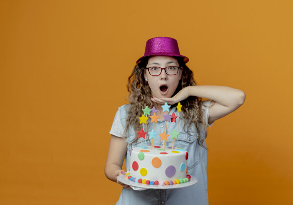女孩惊讶的小女孩戴着眼镜 戴着粉红色的帽子 手里拿着生日蛋糕 把手放在下巴下面 孤立地放在橙色的背景上戴着蛋糕惊喜