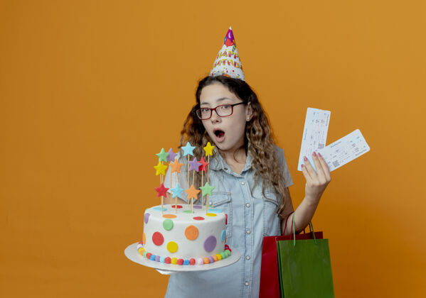 帽子惊讶的年轻女孩戴着眼镜 戴着生日帽 手里拿着生日蛋糕和礼品袋 橙色背景上孤立着女孩礼物包