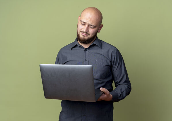不愉快未出租的年轻秃头呼叫中心男子举行 并看着笔记本电脑孤立在橄榄绿的背景与复印空间年轻中心橄榄