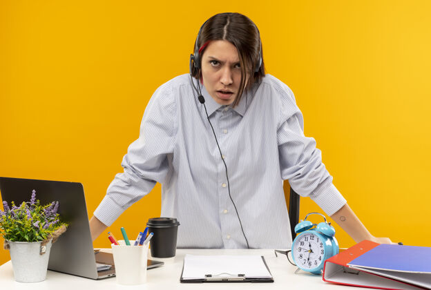 穿着戴着耳机的年轻呼叫中心女孩站在办公桌旁 工作工具被隔离在橙色背景上中心耳机工具