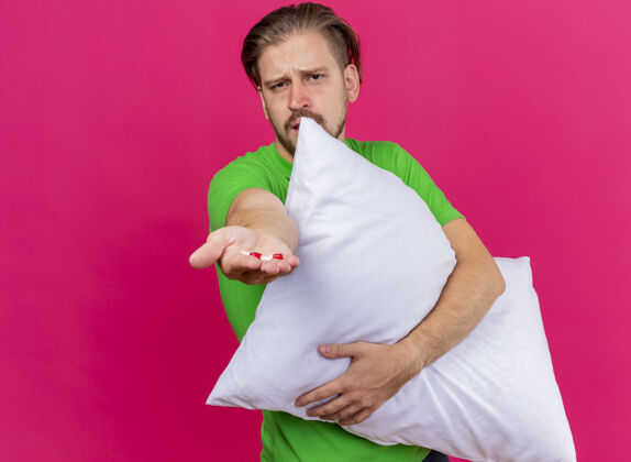 枕头皱眉的年轻英俊的斯拉夫病夫抱着枕头向前伸展胶囊看着前面的粉红色墙壁上孤立的复制空间人拥抱伸展
