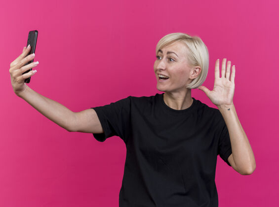 表情快乐的中年金发斯拉夫女人抱着手机 看着手机做着“嗨”的手势孤立在粉红色的墙上衣服姿势电话