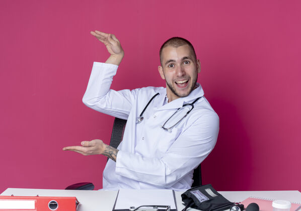 男性令人印象深刻的年轻男性医生穿着医用长袍和听诊器坐在办公桌上与工作工具显示大小与粉红色的手隔离印象听诊器粉色