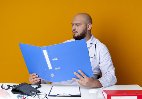 听诊器严格年轻的秃头男医生穿着医用长袍和听诊器坐在工作台上拿着医疗工具 看着橙色背景上孤立的文件夹工具长袍坐着