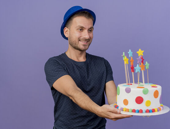 副本戴着蓝色帽子的白人帅哥高兴地拿着生日蛋糕 看着紫色背景上的复制空间帽子拿着生日