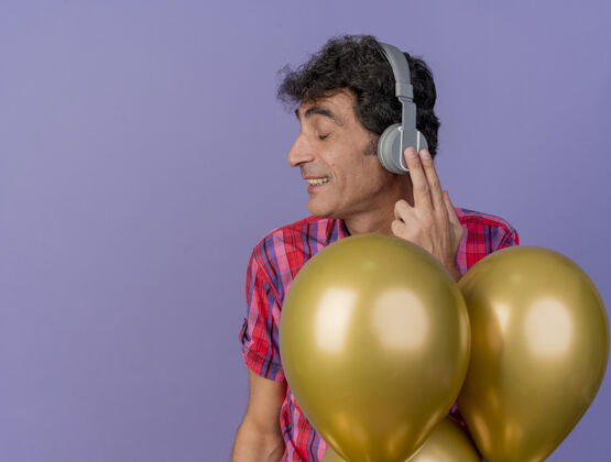 站立微笑的中年白人聚会男子戴着耳机站在气球后面 闭着眼睛触摸耳机 在紫色背景上隔离 留有复印空间触摸紫色派对