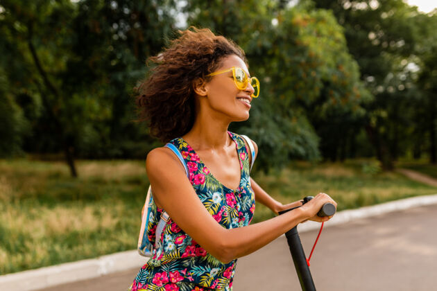 女士年轻时尚的黑人女子在公园里玩得很开心骑着电动脚踏车在夏天的时尚风格 五颜六色的时髦服装 穿着背包和黄色太阳镜微笑道路时尚