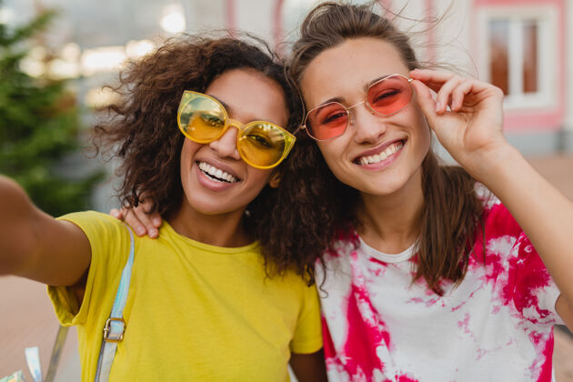 年轻五颜六色的照片 快乐的年轻女孩朋友们微笑着坐在街上用手机自拍 女人们在一起玩得很开心年轻肖像眼镜
