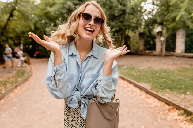微笑迷人的金发美女穿着时髦的服装 戴着优雅的太阳镜和钱包在公园散步休闲女士优雅