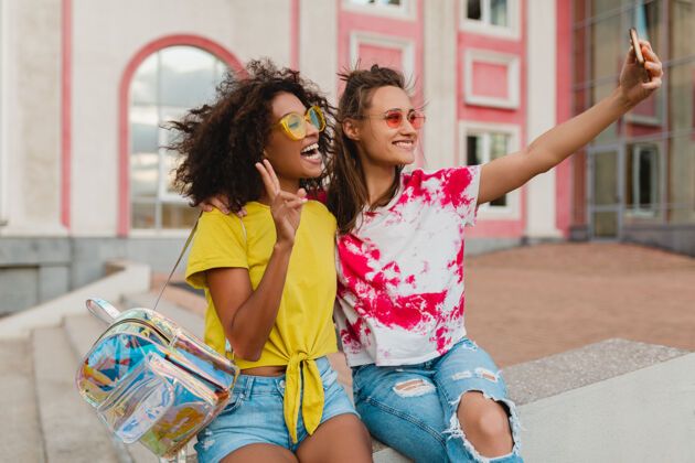 时尚快乐的年轻女孩朋友们微笑着坐在街上用手机自拍 女人们一起开心多种族积极黑色