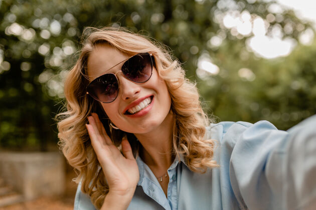 微笑迷人的金发微笑的白牙女人穿着夏装在公园里散步在电话里自拍积极时尚欢乐
