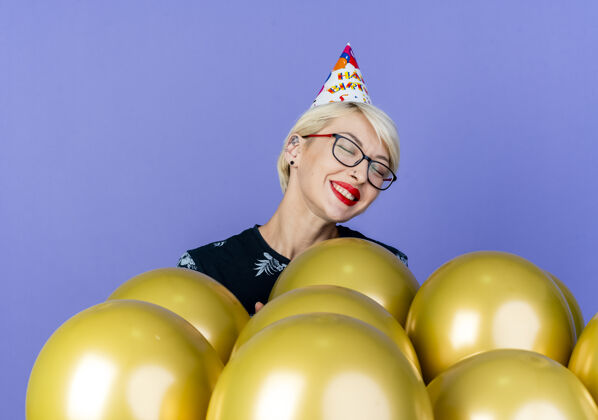 生日微笑的金发派对女孩戴着眼镜和生日帽站在气球后面 闭着眼睛孤立地站在紫色背景上女孩紫色眼镜