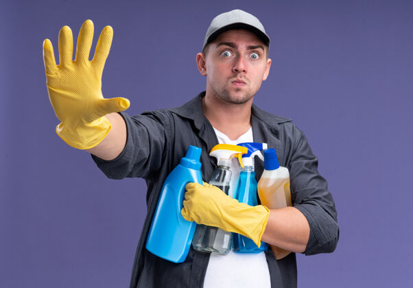 害怕吓坏了年轻帅气的清洁工 戴着t恤 戴着帽子和手套 手里拿着清洁工具 手被隔离在紫色的墙上清洁家伙手套