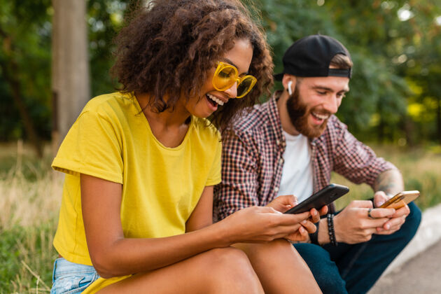 音乐快乐的年轻微笑的朋友坐在公园里用智能手机 男人和女人一起玩多种族情绪微笑