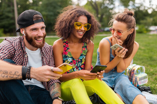 微笑快乐的年轻朋友们坐在公园里用智能手机微笑 男人和女人一起玩耳机情绪友谊