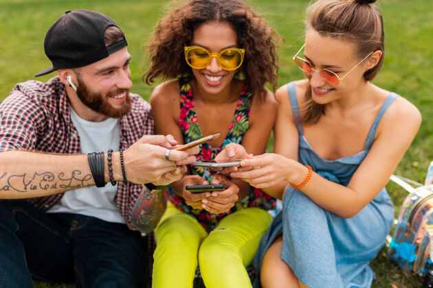 人快乐的年轻朋友们坐在公园里用智能手机微笑 男人和女人一起玩多样性女人年轻
