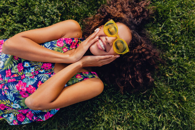 女性年轻时尚的黑人女子戴着无线耳机听音乐在公园里玩得很开心 夏日时尚风格 五颜六色的时髦装扮 躺在草地上 俯瞰风景耳机休闲卷发