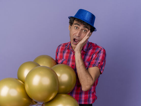 气球惊讶的中年白种人党男子戴着党的帽子拿着气球手放在脸上看着相机孤立的紫色背景与复制空间空间紫色复制