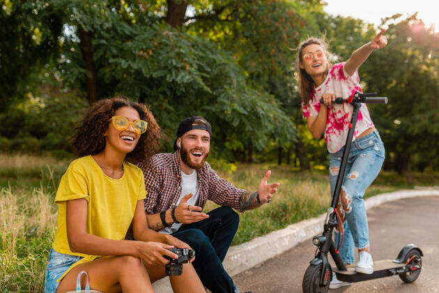 女士快乐的年轻朋友们坐在公园的草地上 带着电动脚踏车微笑着 男女同乐酷女性积极