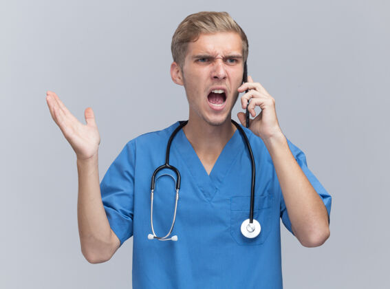 说话愤怒的年轻男医生穿着医生制服 手持听诊器在电话里讲话 手被隔离在白墙上电话医生制服