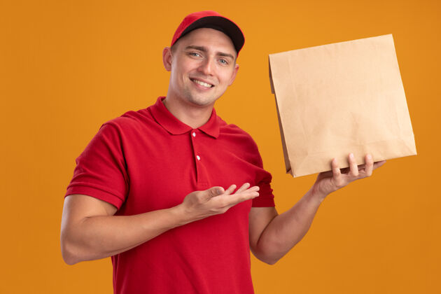男人面带微笑的年轻送货员身穿制服 手持帽子 用手指着隔离在橙色墙上的纸质食品包包装点食物