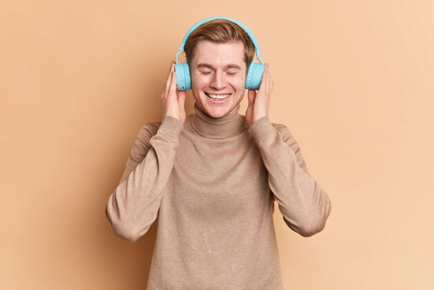 耳机高兴的年轻人闭上眼睛放松 通过无线蓝色耳机收听喜爱的歌曲 使用音乐应用程序微笑 愉快地穿着休闲套头衫姿势促销跳投请