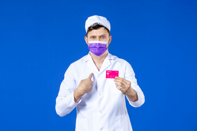 信用前视图中的男医生穿着医疗服和紫色面具持有信用卡在蓝色诉讼病毒医院