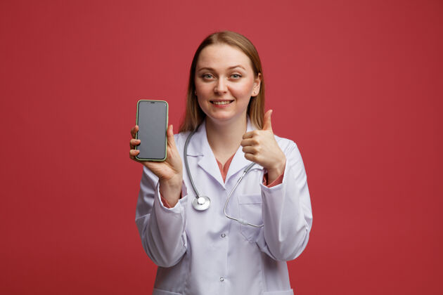 拇指微笑着的年轻金发女医生 穿着医用长袍 脖子上戴着听诊器 手里拿着手机 竖起大拇指脖子听诊器女性