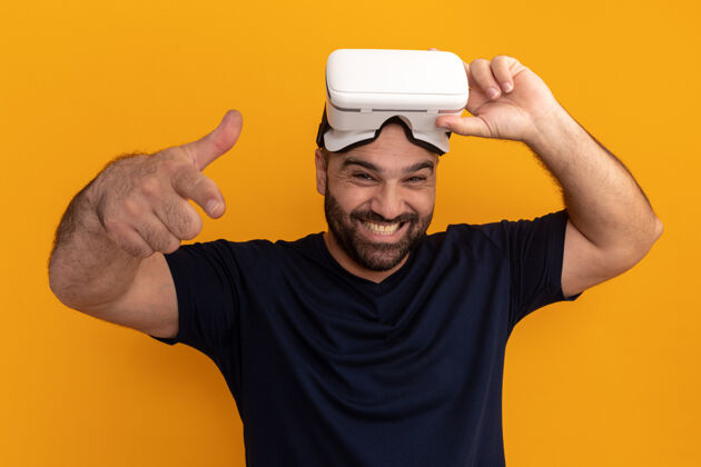 虚拟戴着虚拟现实眼镜 留着海军t恤的大胡子男人快乐而兴奋地用食指指着橙色的墙壁指向胡须现实
