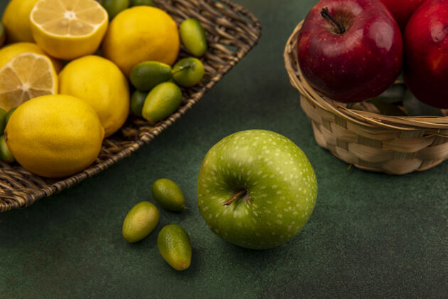 营养柳条托盘上的柑橘类水果柠檬俯视图 绿色表面上有金盏花和绿色苹果厨房苹果切片