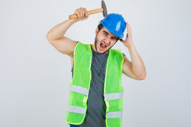 锤子身着制服的年轻建筑工人用锤子敲击头部 看上去很滑稽 正面视图年轻头男性