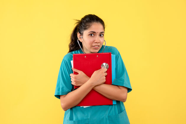 抱着正面图身穿制服的年轻女医生拿着红色的剪贴板站在黄色的背景上前面人快乐