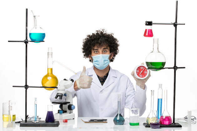 桌子正面图：男药剂师穿着医疗服 戴着面具 白色桌子上放着钟科学前面病毒