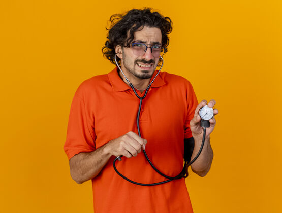 戴戴着眼镜和听诊器的年轻白种人 拿着血压计 隔离在橙色的墙上 留有复印空间疾病血压计橙色