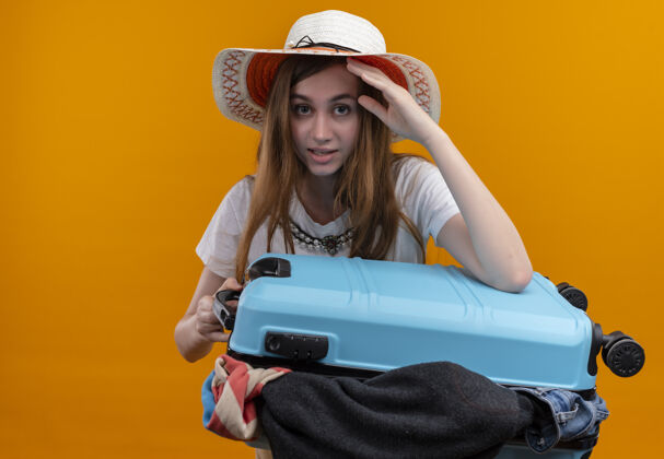 拿着年轻的旅行家女孩戴着帽子 提着装满衣服的手提箱 手放在头上 看着孤立的橘黄色墙壁 还有复制空间满的手提箱女人