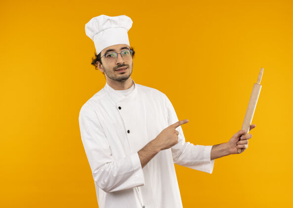 烹饪高兴的年轻男厨师穿着厨师制服 拿着眼镜 指着隔离在黄色墙上的擀面杖卷别针抱