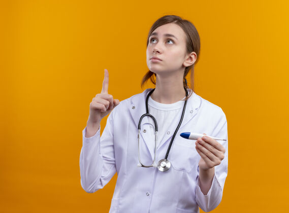 女人体贴的年轻女医生 穿着医用长袍 戴着听诊器 用手指举着温度计 站在隔离的橙色墙上 留着复印空间医生年轻人手指