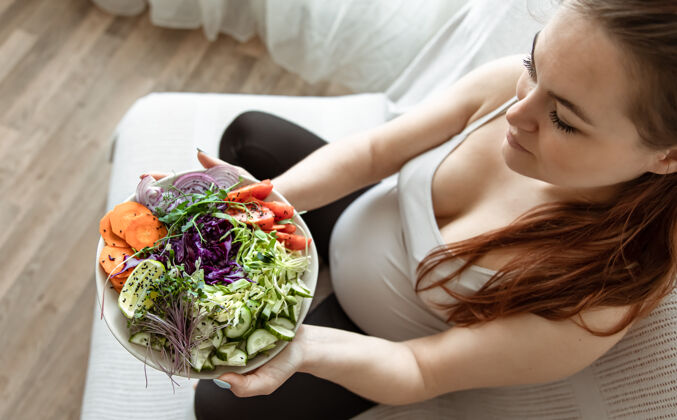 健康孕妇拿着一盘新鲜蔬菜沙拉在家的沙发上俯瞰素食沙拉自然
