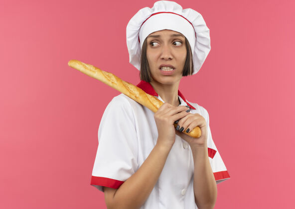 年轻焦急的年轻女厨师穿着厨师制服拿着面包棒看着一旁孤立的粉红色制服烹饪壁板