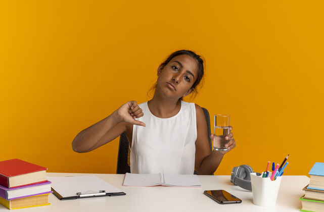 玻璃悲伤的年轻女学生坐在书桌旁 手里拿着一杯水 拇指被孤立地放在橙色的墙上学校女学生趴下