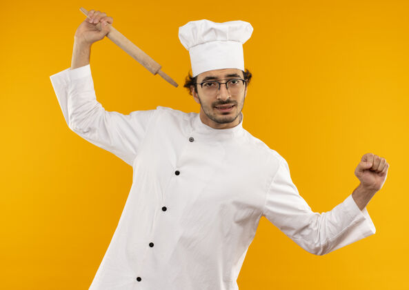 别针疲惫的年轻男厨师穿着厨师制服 戴着眼镜 拿着擀面杖 在黄色的墙上做伸展运动伸厨师抱
