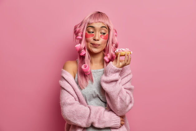 套头衫照片中的亚洲年轻女子看着开胃的甜甜圈 用卷发器做头发 涂上胶原蛋白垫 穿着暖和的毛衣美味面具姿势