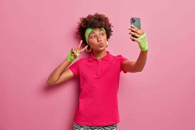 媒体卷发活跃的女人 穿着粉色t恤 头带和运动手套 自拍 做胜利手势 拿着手机 沉迷于社交网络 在室内摆姿势分享风格室内