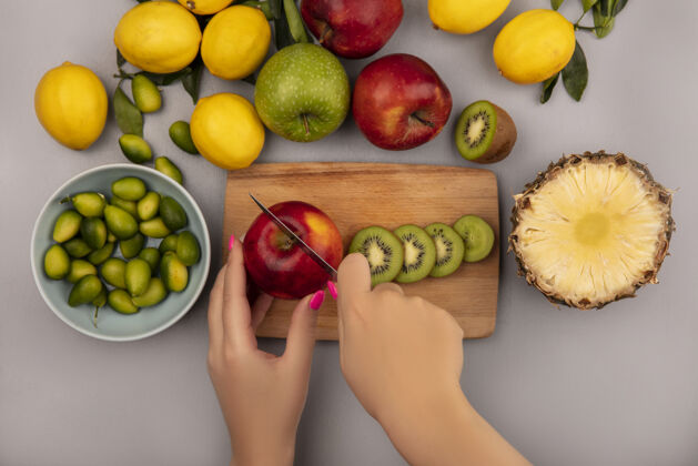 特写俯视图：女性用刀在木制菜板上切苹果 碗上放着金盏花 苹果 奇异果 菠萝和柠檬 隔离在白色的墙上农业刀菠萝