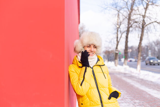 肖像在一个冬天阳光明媚的日子里 一个穿着暖和衣服的红墙背景上的快乐的年轻女子在雪城的人行道上微笑着打电话通信科技城市