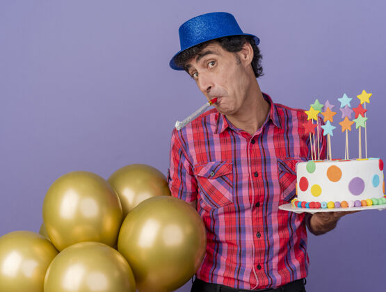 聚会印象深刻的中年派对男子戴着派对帽拿着气球和生日蛋糕吹着派对吹风机看着前面孤立的紫色墙壁脸感情吹