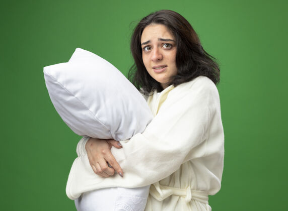 枕头焦急的年轻白种人生病的女孩穿着长袍站在侧视图拥抱枕头看着隔离在绿色背景上的相机女孩视图年轻