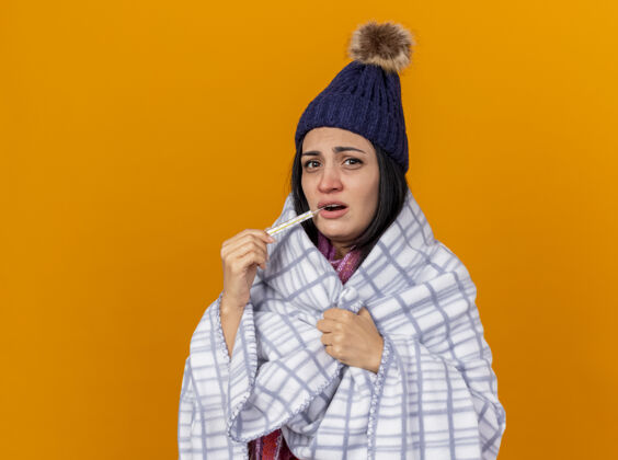 女孩虚弱的年轻白种人生病的女孩戴着冬天的帽子 围巾裹着格子布把温度计放进嘴里抓着格子布隔离在橙色背景上复制空间空间围巾虚弱