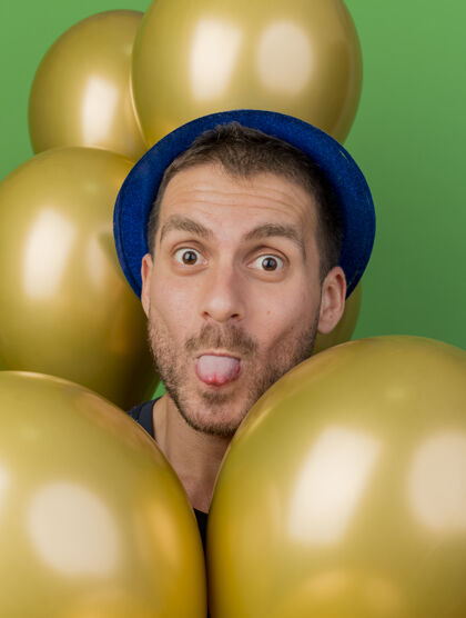 姿势有趣的帅哥戴着蓝色的派对帽 用氦气球在绿色的墙壁上孤立地跺着舌头站着黄金立场人